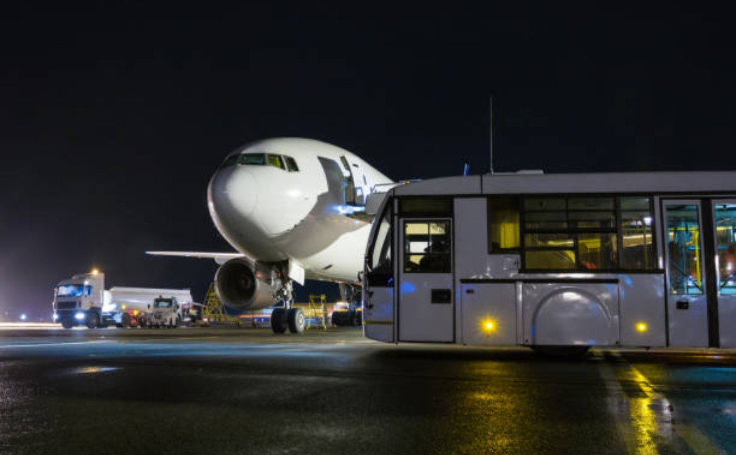 Airport Transportation Services bus parked on the tarmac at night beside a large commercial airplane, with ground support vehicles nearby under runway lights.
