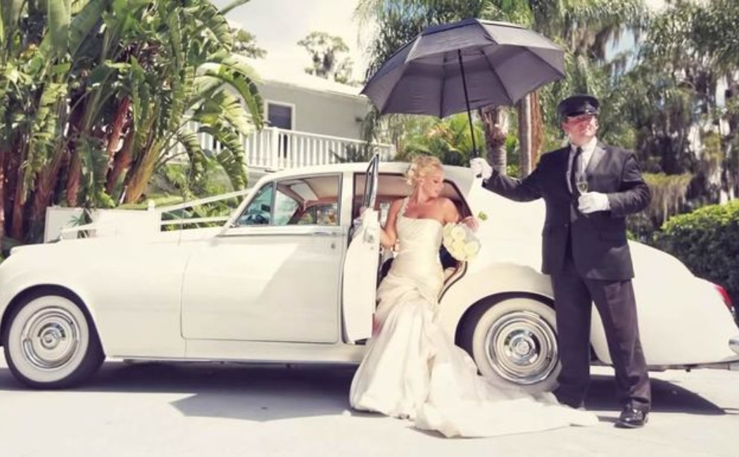 Bride stepping out of a vintage white car while a chauffeur holds an umbrella, highlighting elegant and stylish Wedding Transportation.
