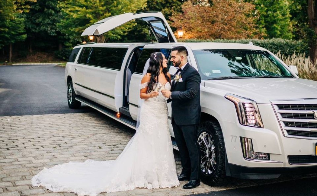Bride and groom standing beside a white stretch limousine, celebrating outdoors on their wedding day. Elegant Wedding Limo Transportation providing luxury arrival for the newlyweds.